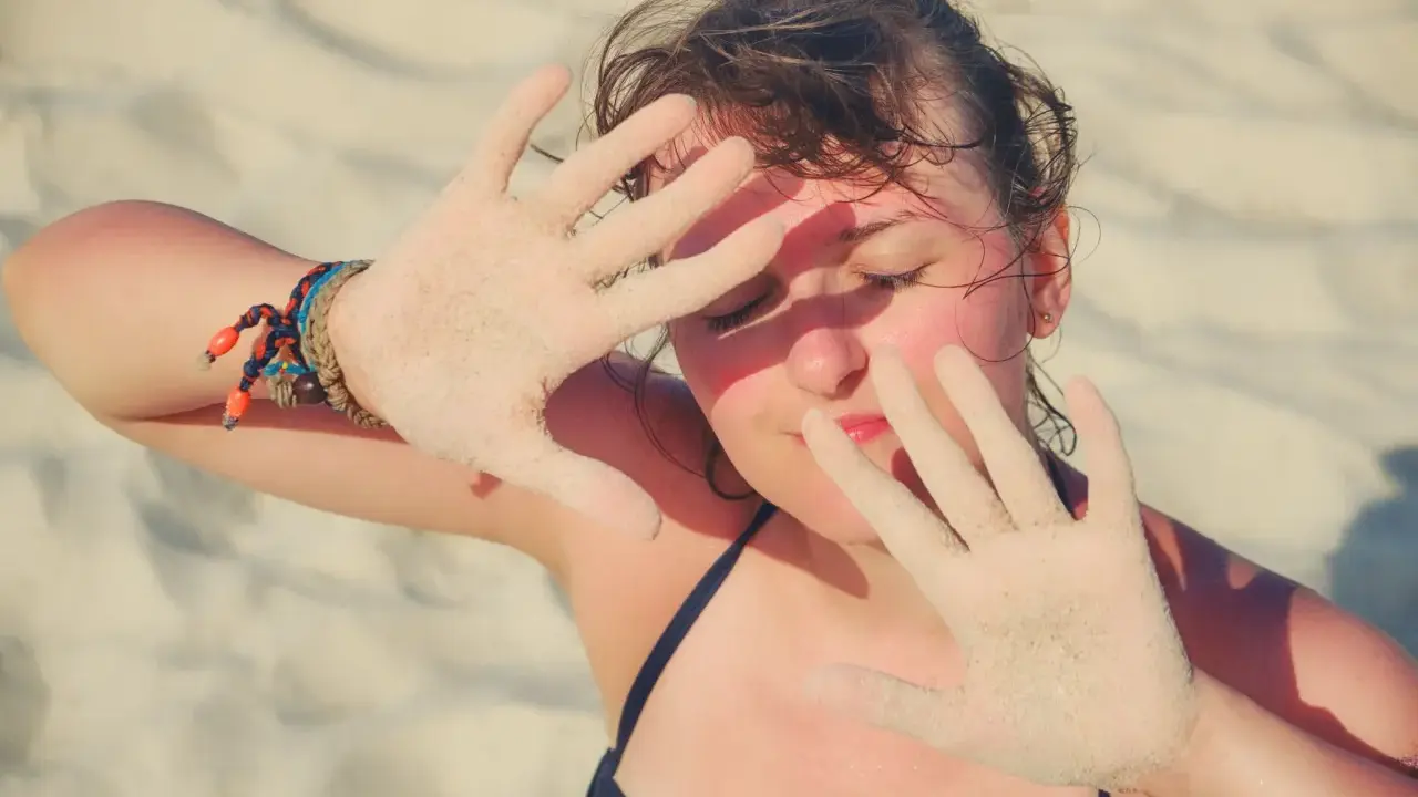 Dziewczyna na plaży zasłania twarz dłońmi pokrytymi piaskiem, być może chroniąc się przed słońcem z powodu fotodermatozy.
