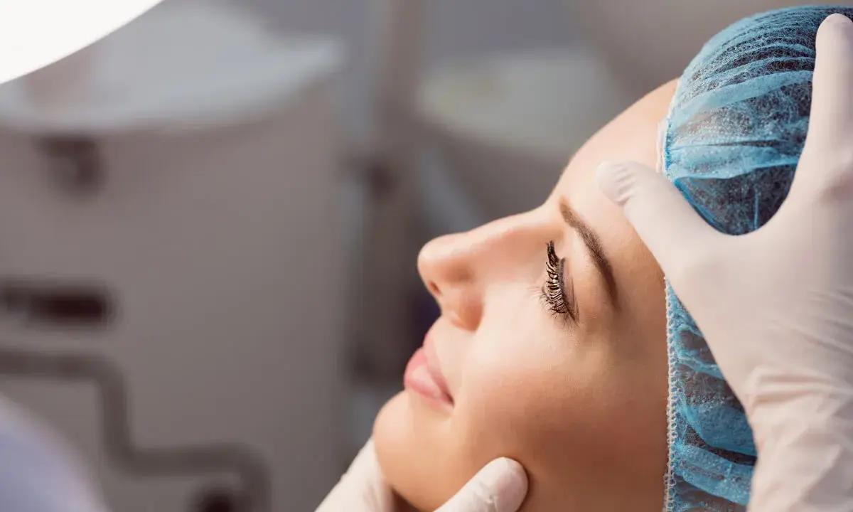 Dermatolog w białej rękawiczce delikatnie bada twarz kobiety w czepku. To wizyta u specjalisty, który pomoże zadbać o zdrową skórę.