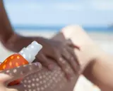 Ochrona przed słońcem na plaży. Ręka aplikuje spray z filtrem UV na nogę, zapewniając skuteczną fotoprotekcję.