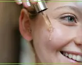Kropla serum z pipety spada na policzek uśmiechniętej kobiety. Dowiedz się, jak stosować retinol dla pięknej cery.