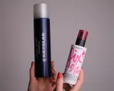 Dwie ręce trzymają dwa spraye: duży, granatowy Kryolan Fixing Spray i mniejszy, biały Fix+ Stay Over MAC. Oba to profesjonalne produkty typu fixer.