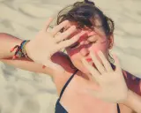 Dziewczyna na plaży zasłania twarz dłońmi pokrytymi piaskiem, być może chroniąc się przed słońcem z powodu fotodermatozy.