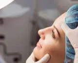 Dermatolog w białej rękawiczce delikatnie bada twarz kobiety w czepku. To wizyta u specjalisty, który pomoże zadbać o zdrową skórę.