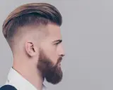Mężczyzna z zarostem i fryzurą slick back, z wygolonymi bokami i zaczesanymi do tyłu włosami.