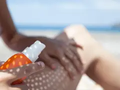 Ochrona przed słońcem na plaży. Ręka aplikuje spray z filtrem UV na nogę, zapewniając skuteczną fotoprotekcję.