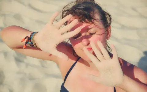 Dziewczyna na plaży zasłania twarz dłońmi pokrytymi piaskiem, być może chroniąc się przed słońcem z powodu fotodermatozy.