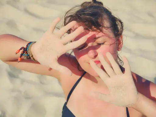 Dziewczyna na plaży zasłania twarz dłońmi pokrytymi piaskiem, być może chroniąc się przed słońcem z powodu fotodermatozy.