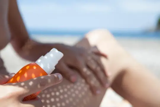 Ochrona przed słońcem na plaży. Ręka aplikuje spray z filtrem UV na nogę, zapewniając skuteczną fotoprotekcję.