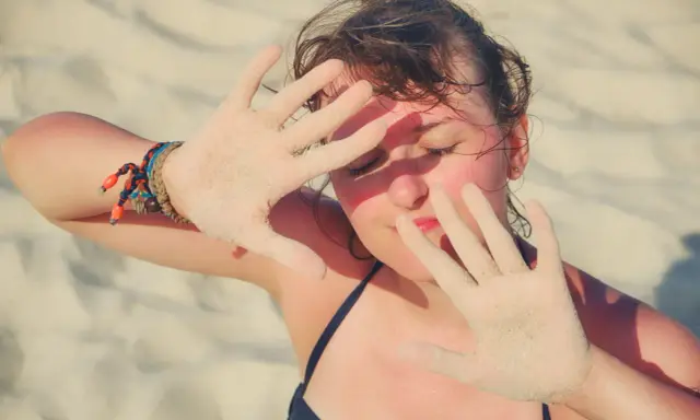 Dziewczyna na plaży zasłania twarz dłońmi pokrytymi piaskiem, być może chroniąc się przed słońcem z powodu fotodermatozy.