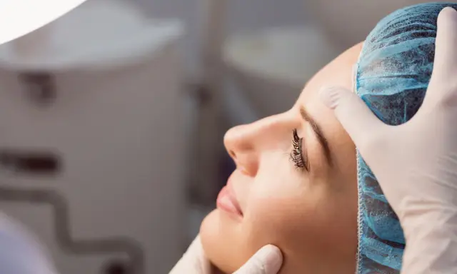 Dermatolog w białej rękawiczce delikatnie bada twarz kobiety w czepku. To wizyta u specjalisty, który pomoże zadbać o zdrową skórę.