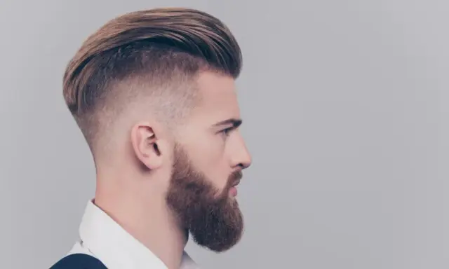 Mężczyzna z zarostem i fryzurą slick back, z wygolonymi bokami i zaczesanymi do tyłu włosami.