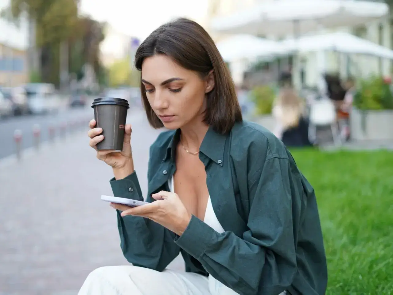 Kobieta o śniadej karnacji z kawą w dłoni przegląda telefon na tle miejskiego krajobrazu.
